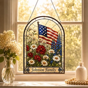 Könnte beinhalten: Buntglasfenster mit der amerikanischen Flagge über einem Feld aus roten, weißen und blauen Blumen. Das Panel ist oben gewölbt und trägt unten den Text "Johnson Family". Es hängt vor einem Fenster.