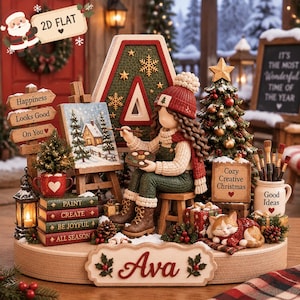 Puede incluir: Un conjunto decorativo de figuras navideñas que muestra a una persona pintando un paisaje invernal en un caballete, rodeada de un árbol decorado, un gato durmiendo, libros apilados con las palabras Paint, Create, Be Joyful, All Season y un letrero que dice Ava.