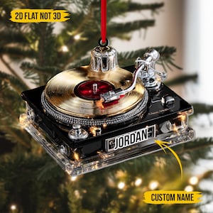 Könnte beinhalten: Schwarz-goldener Weihnachtsbaumschmuck in Form eines Plattenspielers mit dem Namen "JORDAN" auf der Vorderseite. Der Schmuck hat ein rotes Band und den Text "2D FLAT NOT 3D".