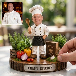 May include: A custom chef figurine on a wooden base labeled CHEF'S KITCHEN, featuring a white uniform, black apron, and chef hat. The display includes a wooden spoon, a small recipe book, fresh herbs, red and yellow peppers, and an onion.