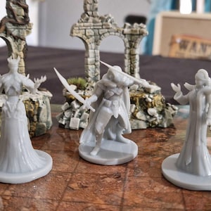 Op de afbeelding: Een tafelscène met miniatuur fantasyfiguren en architecturale elementen. Drie grijze beeldjes van een koningin, een krijger en een tovenares zijn op een spelbord geplaatst. Stenen bogen en een "WANTED"-bord staan op de achtergrond.