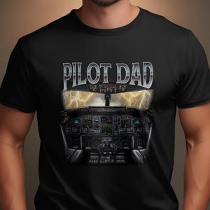 May include: Black t-shirt featuring a detailed illustration of an airplane cockpit with the text "PILOT DAD" in silver lettering above. The cockpit illustration includes gauges, screens, and a view of lightning. A great gift for a pilot.
