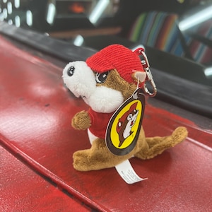 May include: A plush beaver keychain with a red cap and shirt. The beaver has brown fur, a white face, and a tag with a logo. The keychain is attached to a silver metal clasp.