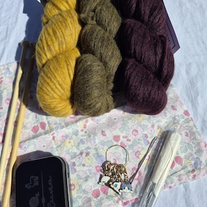 Könnte beinhalten: Drei Garnstränge in Senfgelb, Olivgrün und Dunkelpflaume. Enthalten sind zwei Stricknadeln aus Holz, eine schwarze Metalldose mit Tier-Symbolen und dem Text "soluna", ein Set mit Katzen-Maschenmarkierern sowie eine Häkelnadel aus Kunststoff.
