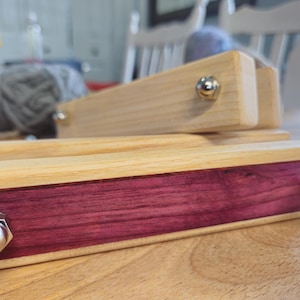 Pode incluir: Ferramentas de tecelagem de madeira com acabamento natural e uma incrustação de madeira vermelho-púrpura contrastante. As ferramentas são presas por parafusos prateados. Ferramentas para arte têxtil e artesanato.