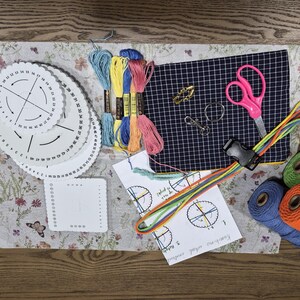 May include: A kumihimo craft kit featuring white circular and square braiding disks, colorful embroidery floss, a navy blue checkered fabric square, pink scissors, metal clasps, a black plastic buckle, instruction sheets, and spools of cord.