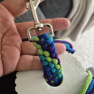 May include: A braided leash attachment with a silver metal clasp. The braided section features a pattern of blue, green, and purple threads. The leash is attached to a white, scalloped-edged object.