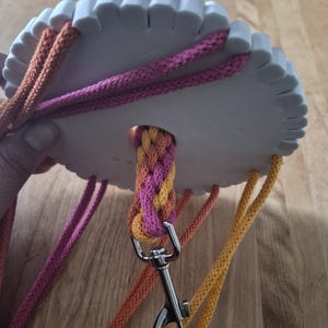 Può includere: Un disco per intrecciare kumihimo circolare bianco con bordi dentellati, caratterizzato da molteplici fili di corda rosa, arancione e gialla. Una sezione parzialmente intrecciata della corda passa attraverso il foro centrale, fissata a un moschettone girevole in metallo argentato.