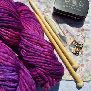 May include: A skein of thick, hand-dyed yarn in shades of magenta, purple, and violet. Included are two light-colored wooden knitting needles, a white crochet hook, a small black metal tin labeled "soluna" with animal icons, and a gold stitch marker.