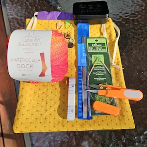 Pode incluir: Uma coleção de materiais de tricô em tecido amarelo. Inclui um novelo de fio de meia aquarela, agulhas de tricô, uma fita métrica, pequenas tesouras laranja e uma lata preta. O rótulo do fio diz "LAINES du NORD WATERCOLOR SOCK".