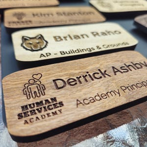 May include: Wooden name badges with engraved text. One badge reads "Derrick Ashby, Academy Principal" with the Human Services Academy logo. Another reads "Brian Raho, AP - Buildings & Grounds" with a fox logo. Other badges are visible.