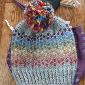 May include: A gray knit hat with a rainbow chevron pattern and a large, multicolored pom-pom. The hat is handmade and features a ribbed brim. Knitting needles and a small coaster with text are also visible.