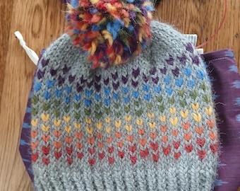 Könnte beinhalten: Graue Strickmütze mit Regenbogen-Zackenmuster und einem großen, mehrfarbigen Bommel. Die Mütze ist handgefertigt und hat einen gerippten Bund. Stricknadeln und ein kleiner Untersetzer mit Text sind ebenfalls sichtbar.