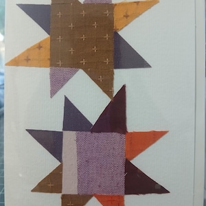 May include: Two star-shaped quilt blocks on a white background. The stars are made of fabric in shades of brown, orange, purple, and lavender. The center of the top star is brown with small cross-shaped designs. The bottom star has a lavender center.