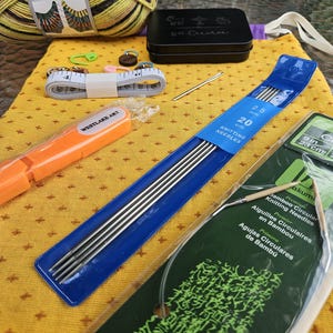 May include: A collection of knitting supplies on a yellow patterned fabric. Includes yarn balls, a black tin, a measuring tape, knitting needles (2.5 mm, 20 cm), and a circular knitting needle package.