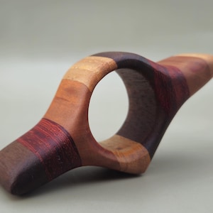 May include: A handcrafted wooden book holder with a unique, ergonomic design. The holder features a variety of wood tones, including light brown, dark brown, and reddish-brown stripes. The curved shape allows for easy page turning and comfortable reading.