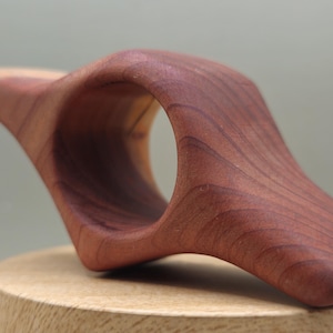 May include: A handcrafted wooden book holder in a rich, reddish-brown hue. The ergonomic design features a curved shape with a central opening, ideal for holding a book open. The wood grain is visible, adding to its natural appeal.
