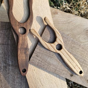 May include: Two wooden yarn tools, one dark brown and one light brown, are displayed on wooden planks. Each tool has a U-shaped top with a circular hole and a hole at the base of the handle. The tools are for fiber arts.