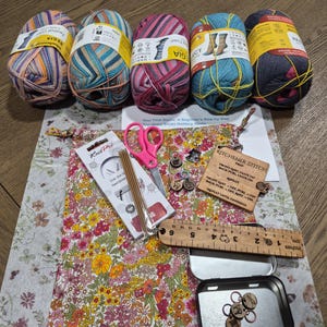 May include: A collection of knitting supplies including five colorful yarn skeins, a floral fabric pouch, pink scissors, knitting needles, wooden stitch markers, a wooden ruler, and a metal tin. The image also includes a beginner's guide to knitting socks.