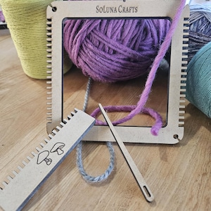 May include: A wooden loom with a purple yarn ball, a gray yarn, a yellow yarn, a green yarn, and a wooden needle. The loom has the text "Soluna Crafts" printed on it.
