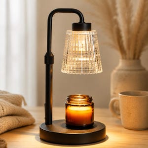 May include: A black metal candle warmer lamp with a curved arm and a circular base. The lamp features a clear, textured glass shade that emits a warm glow, positioned directly above a small amber glass jar containing a lit candle with a visible flame.