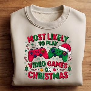 May include: Beige sweatshirt with a Christmas-themed graphic. The graphic features two game controllers, one red and one green, with the text "MOST LIKELY TO PLAY VIDEO GAMES ON CHRISTMAS" in red and green. Festive elements like candy canes and a Santa hat are included.