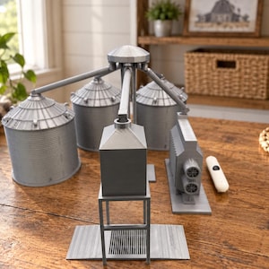 May include: A gray 3D-printed model of a grain elevator complex featuring three cylindrical silos with ribbed textures, a central square tower on a raised frame, and a rectangular building with two circular vents, all connected by overhead pipes.