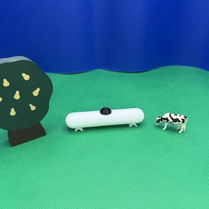 May include: A white toy propane tank with a black top, a green toy tree with yellow pears, and a black and white toy cow on a green surface.