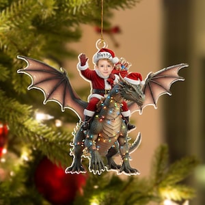 May include: A Christmas ornament featuring a child dressed as Santa Claus riding a dragon. The dragon is adorned with string lights. The child's Santa hat has the name "Harry" on it. The dragon has large wings and is green and gray.