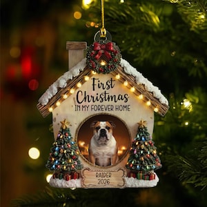 May include: A Christmas ornament shaped like a house, featuring a bulldog's portrait. The ornament has a festive design with a wreath, Christmas trees, and the text "First Christmas in My Forever Home." The dog's name, "Raider," and the year "2026" are also displayed.