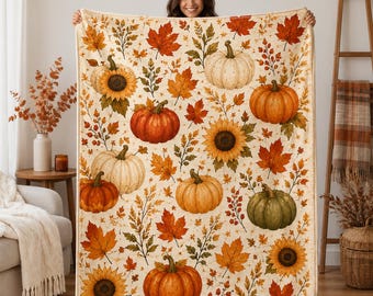 May include: A rectangular throw blanket featuring a repeating pattern of orange, white, and green pumpkins, yellow sunflowers, and autumn leaves in shades of red, orange, and gold on a cream-colored background with a finished stitched edge.