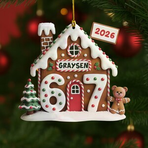May include: A gingerbread house Christmas ornament with white icing, red and green accents, and the name "Graysen." The ornament features the numbers "607" and a small gingerbread bear. A tag displays "2026."