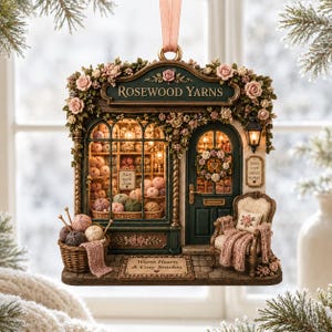 May include: A decorative hanging ornament shaped like a miniature yarn shop named Rosewood Yarns. The facade features floral accents, a display window filled with yarn balls, a dark green door, a small armchair with a blanket, and text signs.