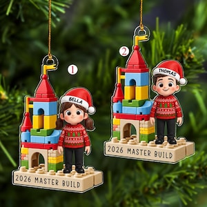 May include: Two flat acrylic Christmas ornaments featuring cartoon characters standing next to colorful building block castles. One ornament shows a character named Bella, the other Thomas. Both wear red sweaters and hats with 2026 MASTER BUILD text.