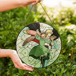 May include: Round stained glass window art featuring a scene with two animated characters. The artwork depicts a woman in a green dress and a man with black wings. The background is light green with floral accents, and the piece is held by a chain.
