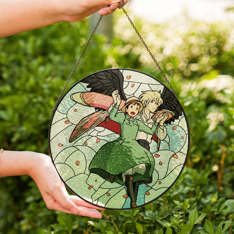 May include: Round stained glass window art featuring a scene with two animated characters. The artwork depicts a woman in a green dress and a man with black wings. The background is light green with floral accents, and the piece is held by a chain.