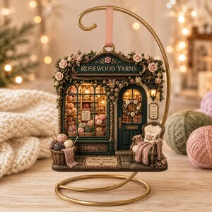May include: A miniature Rosewood Yarns shop ornament hanging from a gold metal stand. The detailed storefront features shelves of yarn, a dark green door, a small armchair with a blanket, and floral accents. Text reads: Rosewood Yarns, Knit with Love, and Warm Hearts & Cozy Stitches.