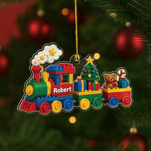 May include: A flat, acrylic Christmas tree ornament shaped like a colorful toy train built from interlocking plastic bricks. The train features a green engine with the name Robert printed in white, a car carrying a decorated tree, and a caboose with a teddy bear and candy cane.