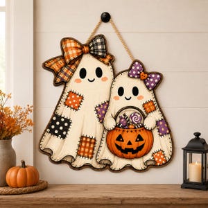 May include: A decorative wall hanging featuring two cartoon ghosts with stitched patchwork patterns in orange, black, and purple. The larger ghost wears a plaid bow, while the smaller ghost holds a jack-o'-lantern bucket filled with candy.