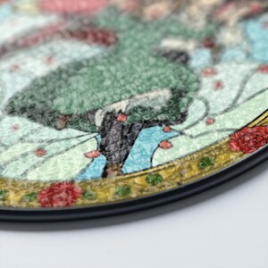May include: A round decorative item with a black border. The design features a figure in a green dress, surrounded by red flowers and a light blue background. The item has a textured, mosaic-like appearance.