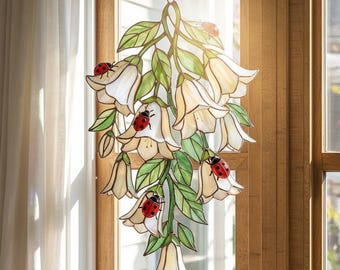 May include: A stained glass hanging decoration featuring white bell-shaped flowers, green leaves, and red ladybugs. The floral arrangement is suspended from a chain and hangs in front of a window. The piece is illuminated by sunlight.