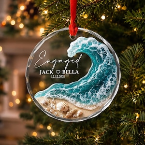 May include: A round clear acrylic ornament featuring a textured resin ocean wave crashing onto a sandy beach with small seashells. The text reads Engaged, JACK HEART BELLA, and 12.12.2026. A red ribbon is attached to the top for hanging purposes.