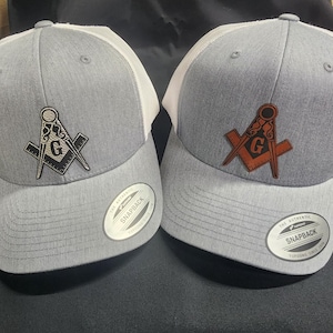 Puede incluir: Dos gorras de camionero grises y blancas con una parte trasera de malla blanca. Ambas gorras presentan un símbolo masónico bordado en blanco y negro en la parte delantera. Una gorra tiene un símbolo bordado marrón. Las gorras tienen una etiqueta negra "Snapback" en la parte trasera.