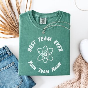 May include: A folded green t-shirt with the text "BEST TEAM EVER" and a graphic of an atom. A silver necklace with a circular pendant rests on the shirt. Blue jeans and a white shoe are also visible.