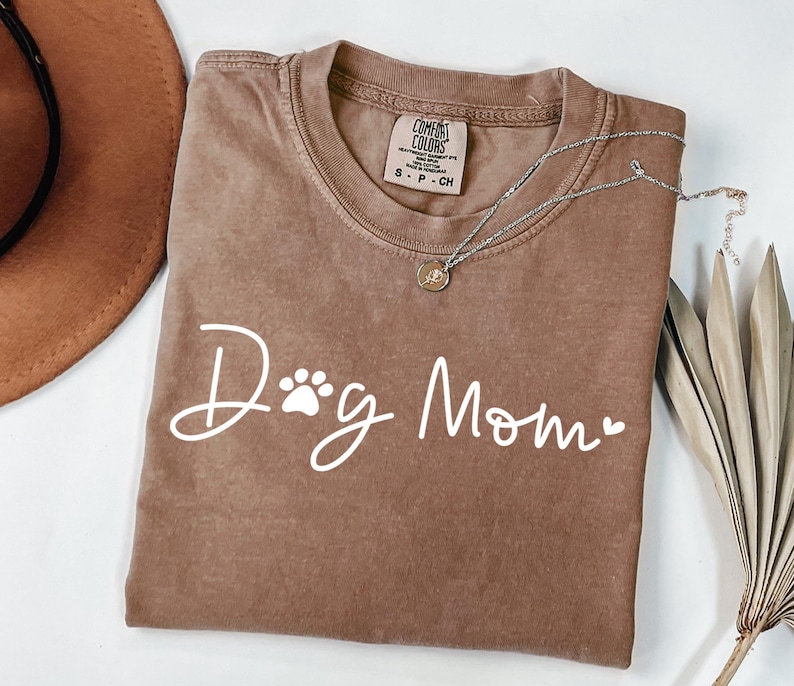 Może przedstawiać: Brązowa koszulka Comfort Colors z białym napisem "Dog Mom", z odciskiem łapy i sercem. Srebrny naszyjnik z wisiorkiem jest zarzucony na koszulkę. Koszulka jest złożona na białej powierzchni.