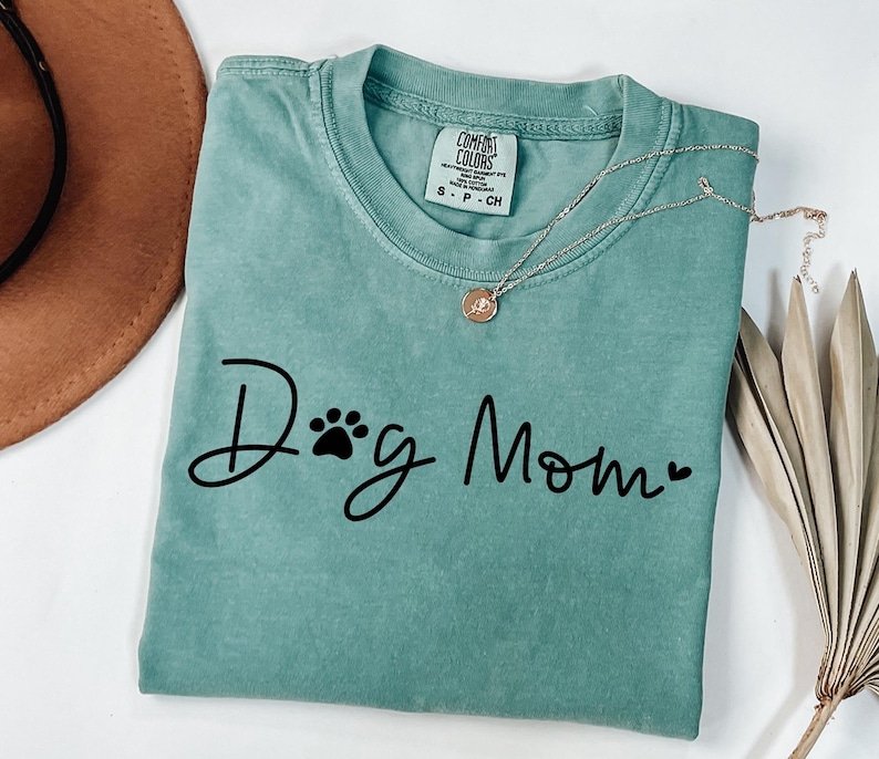 Może przedstawiać: Miętowa koszulka z napisem "Dog Mom" w czarnym skrypcie, z odciskiem łapy i sercem. Srebrny naszyjnik z wisiorkiem spoczywa na koszulce. Koszulka jest złożona, a brązowy kapelusz jest widoczny w tle.