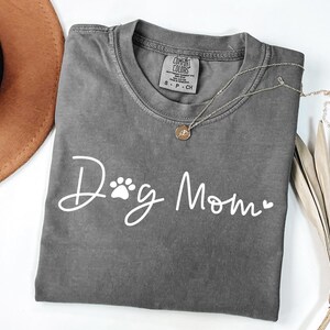 Może przedstawiać: Szara koszulka Comfort Colors z napisem "Dog Mom" w białym skrypcie, z odciskiem łapy i sercem. Złoty naszyjnik z wisiorkiem spoczywa na koszulce. Koszulka jest złożona, a brązowy kapelusz jest widoczny w tle.