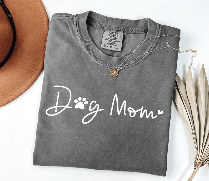 Może przedstawiać: Szara koszulka Comfort Colors z napisem "Dog Mom" w białym skrypcie, z odciskiem łapy i sercem. Złoty naszyjnik z wisiorkiem spoczywa na koszulce. Koszulka jest złożona, a brązowy kapelusz jest widoczny w tle.