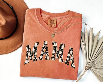 May include: A coral-colored Comfort Colors sweatshirt with the word "MAMA" in a black and white ghost and floral pattern. A gold necklace and a brown hat are also visible.