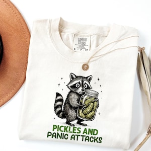 May include: Cream-colored t-shirt featuring a cartoon raccoon clutching a jar of pickles. The raccoon has a worried expression. The text "PICKLES AND PANIC ATTACKS" is printed in green below the raccoon.